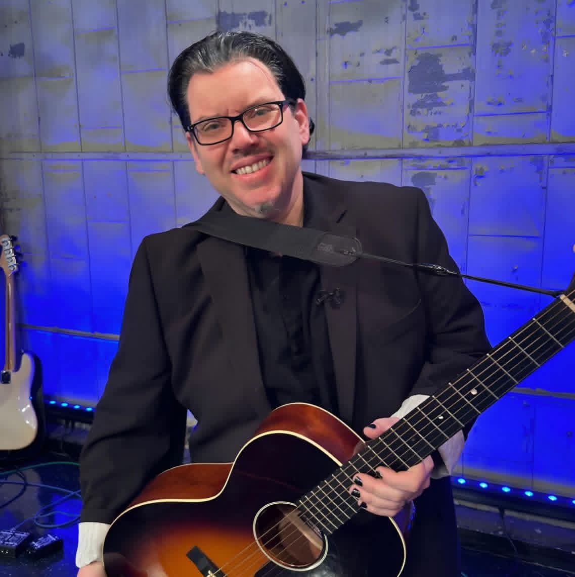 Jack Miele smiling on stage under blue lights, holding a sunburst acoustic guitar