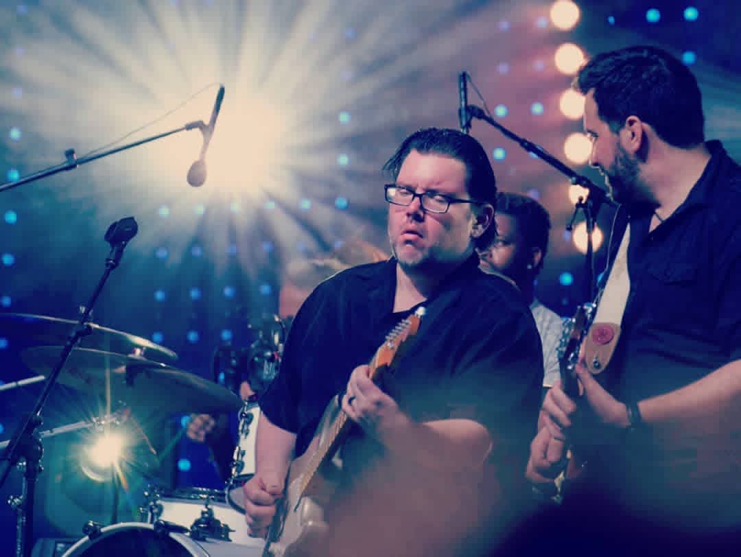Jack Miele on stage playing electric guitar under a bright spotlight with his band