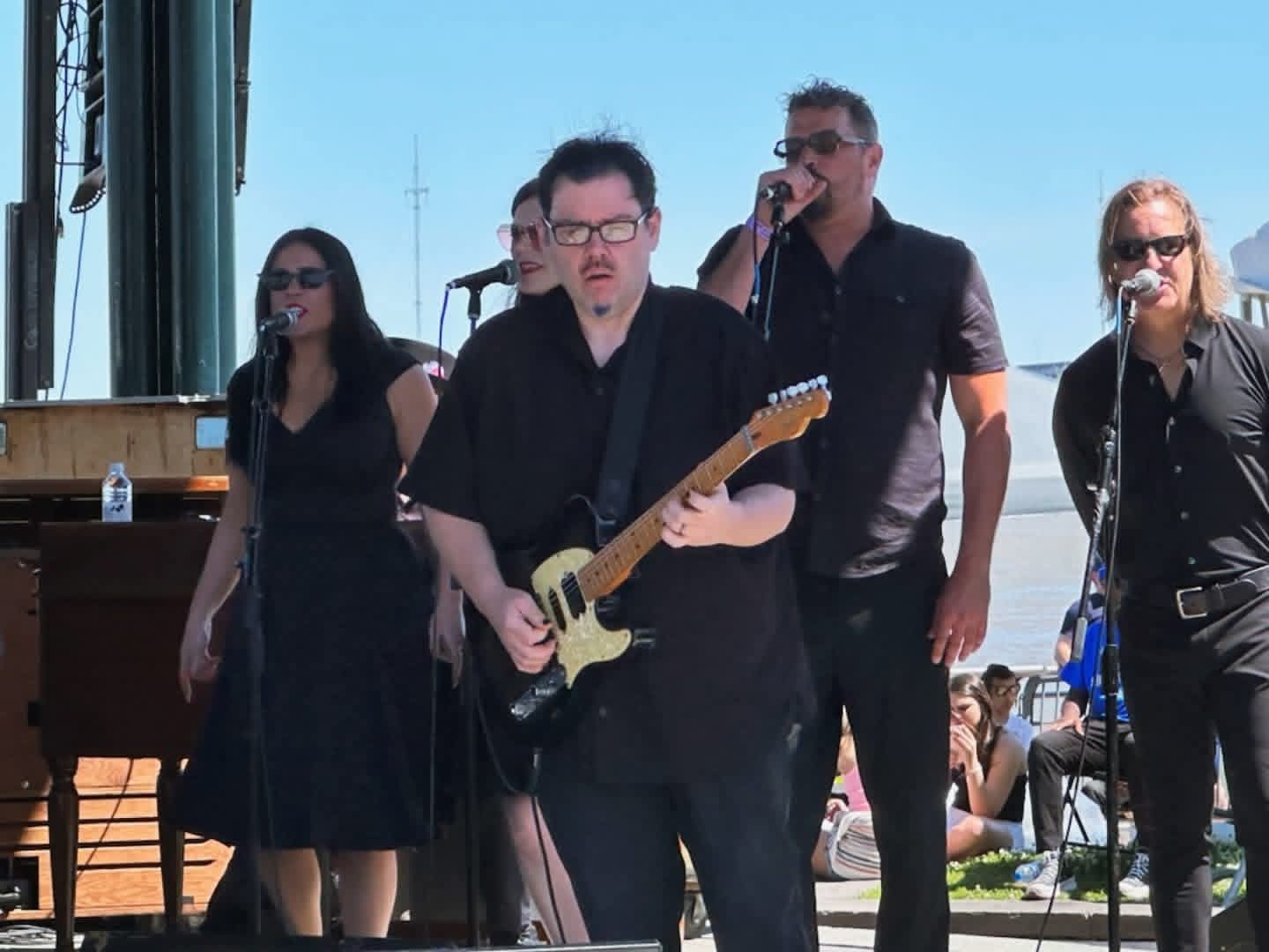 Jack Miele on an outdoor daytime stage playing a black electric guitar with a singer and band