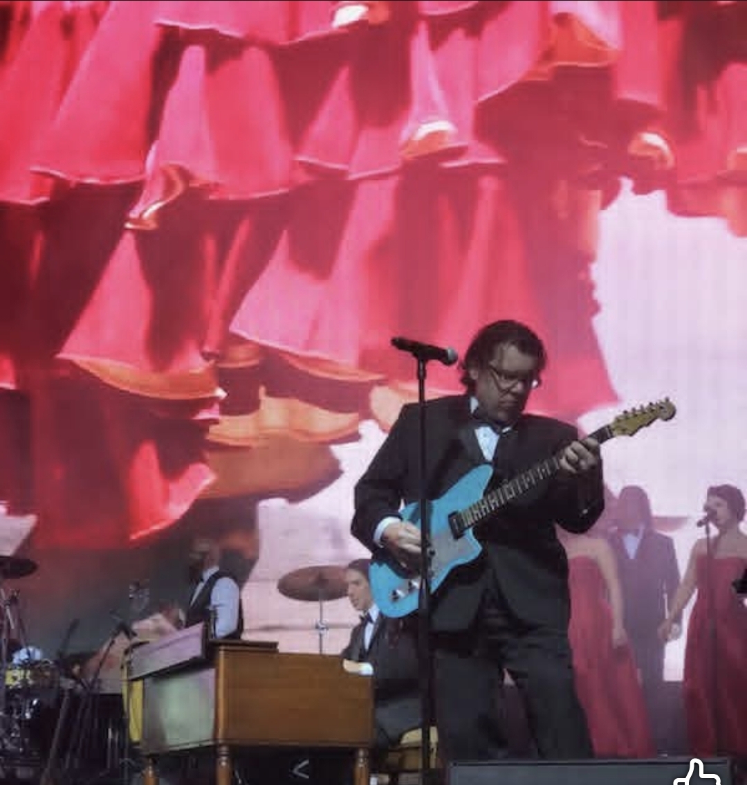 Jack Miele in a suit and bowtie playing a light blue guitar on stage with a red backdrop