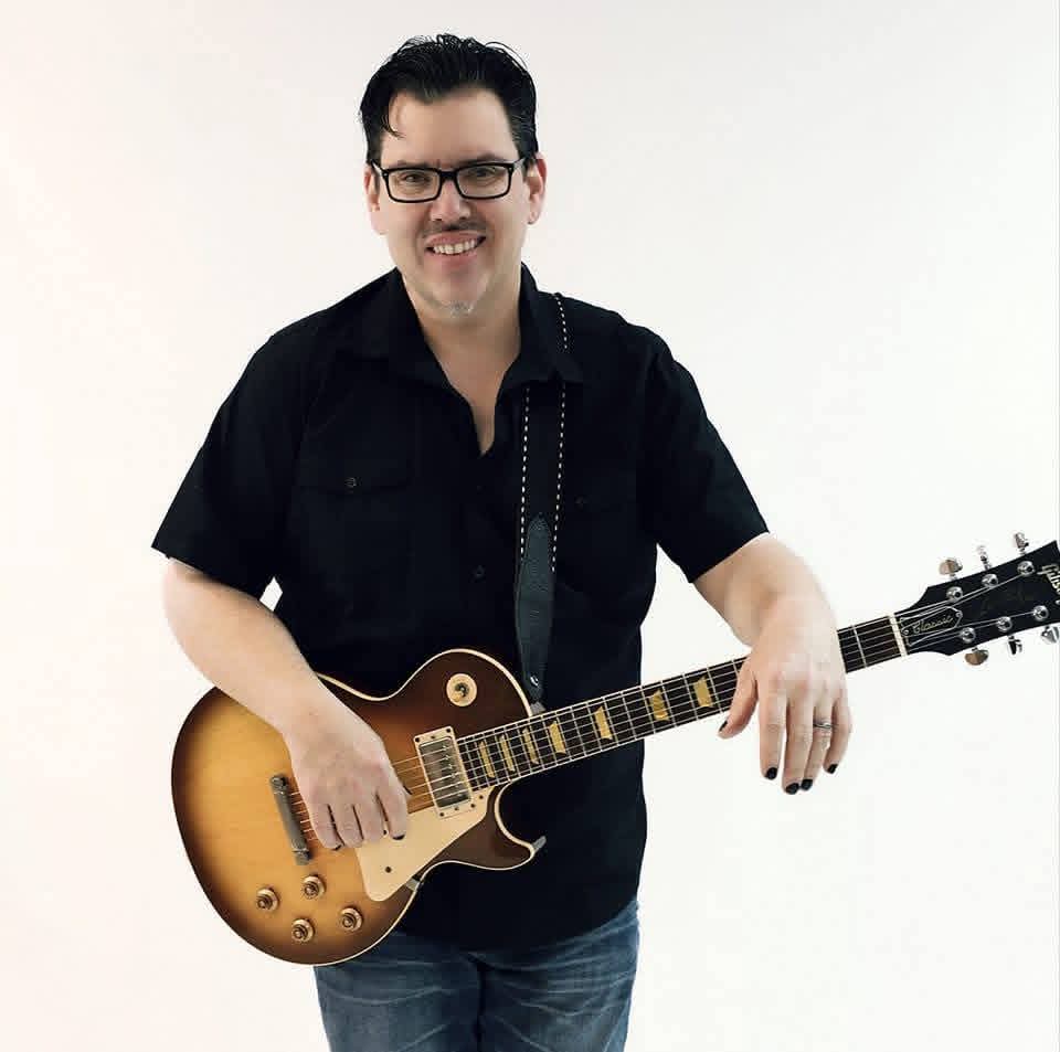 Jack Miele smiling in a promo portrait with a sunburst Gibson Les Paul against a white backdrop