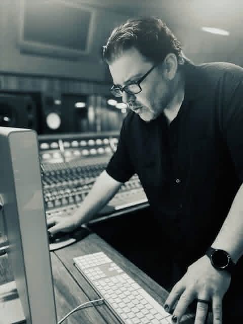 Black-and-white photo of Jack Miele at a keyboard and mouse with a mixing console behind him