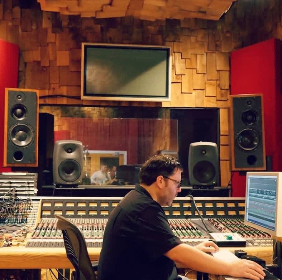Jack Miele seated at a large mixing console with a view into the live room through the control-room window