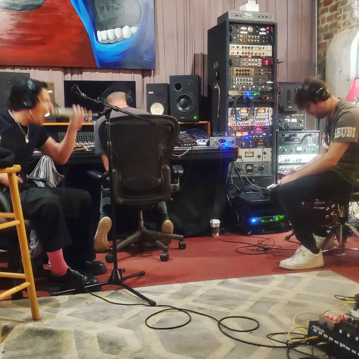 Wide shot of a studio session — vocalist at the mic, engineer at the console, and collaborator on headphones